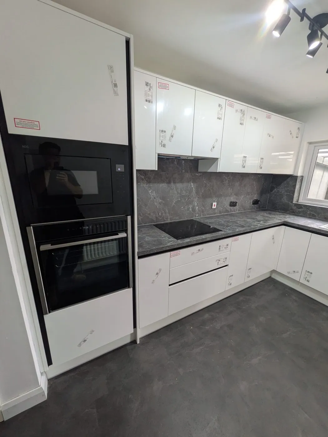 White gloss kitchen with a built-in oven tower and dark stone-effect splashback, protective film still on the doors
